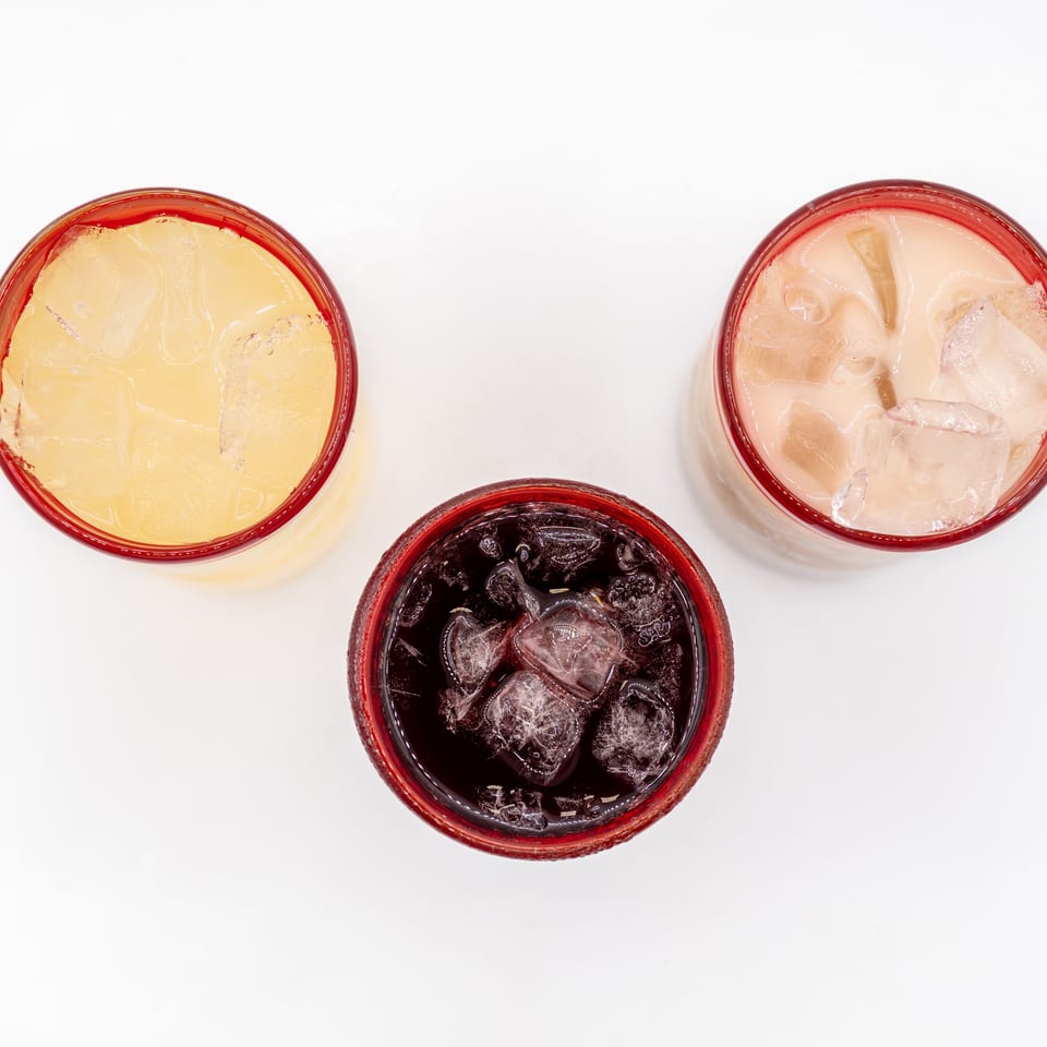 Three red-rimmed glasses of iced beverages—pale yellow, creamy pink, and dark purple—arranged on a white surface.
