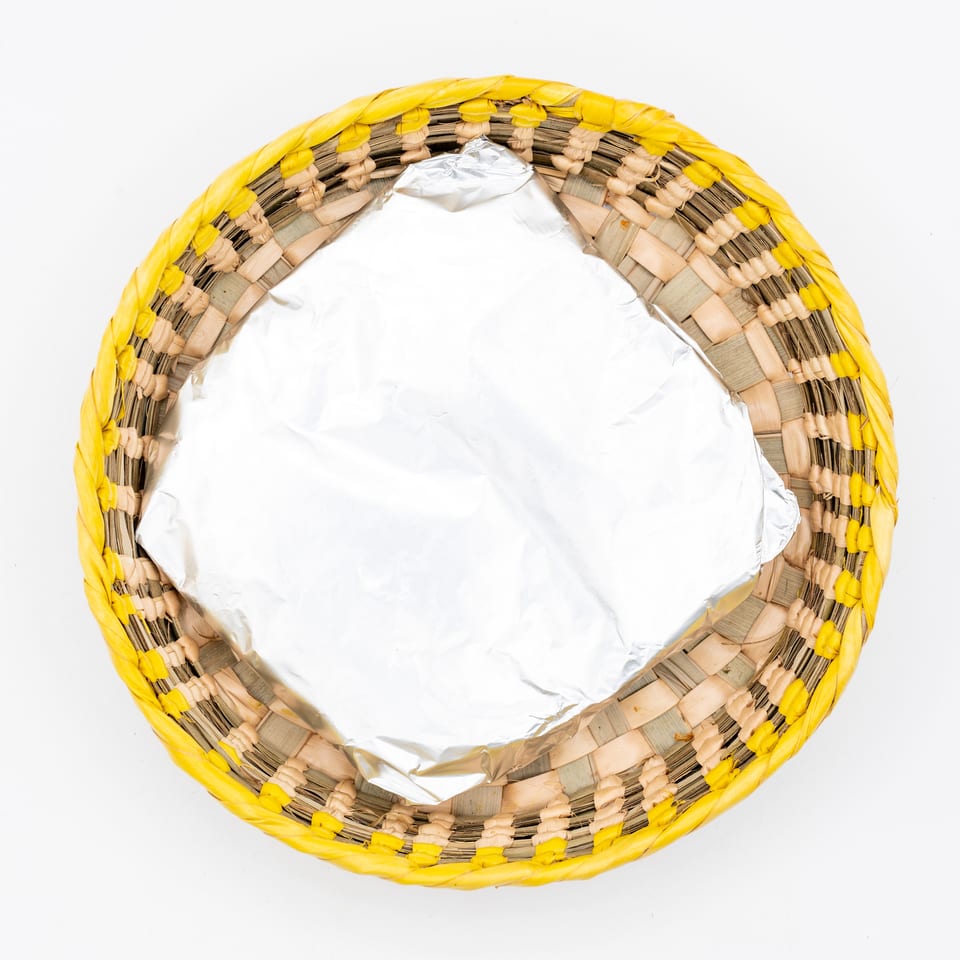 Top-down view of a woven basket with a bright yellow rim, containing a foil-covered dish on a white background.