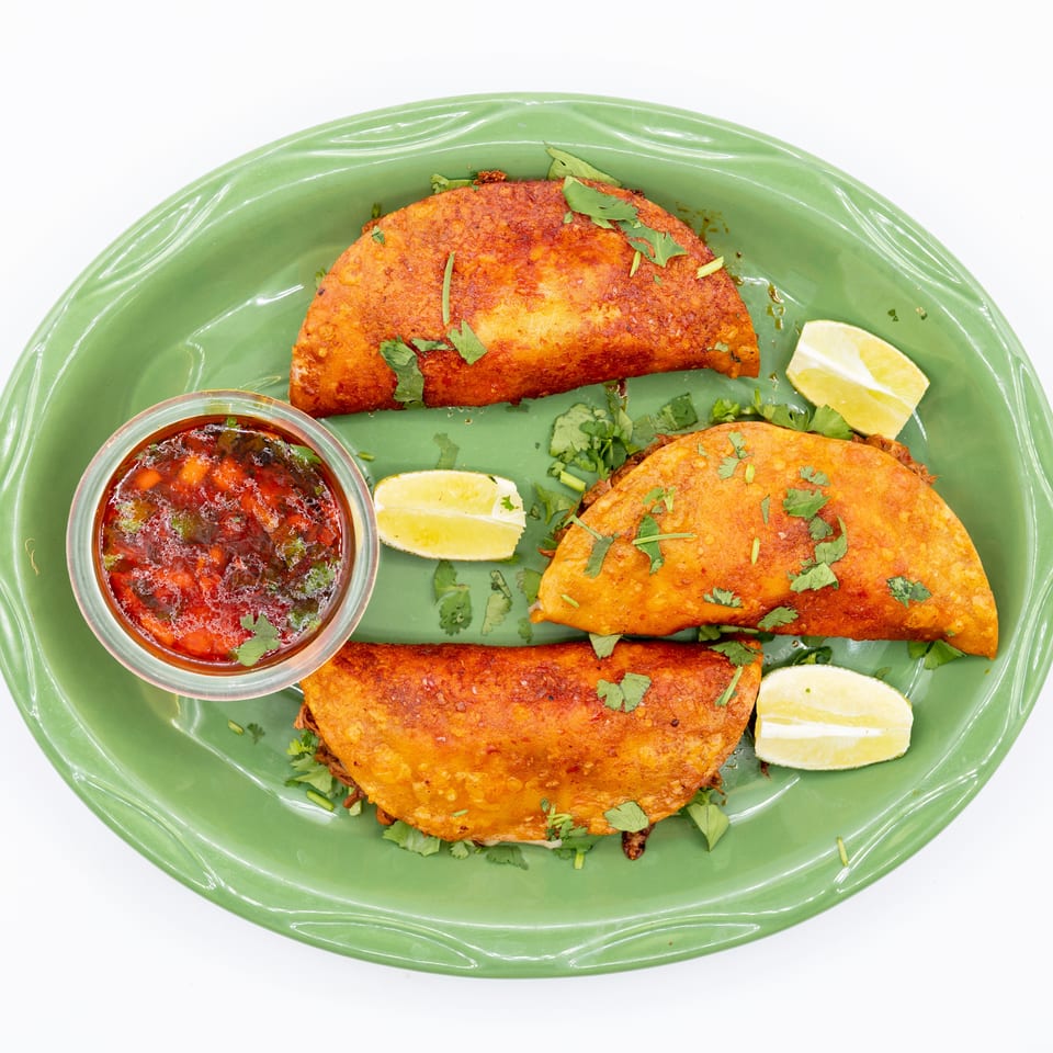 Crispy golden tacos garnished with cilantro, lime wedges, and salsa on a green platter.