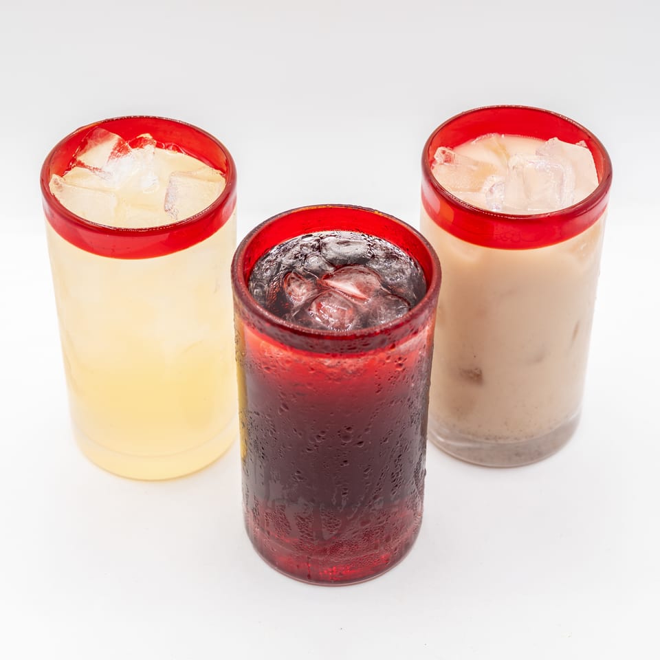 Three red-rimmed glasses of iced beverages, including pale yellow, creamy white, and dark red drinks, arranged on a white surface.