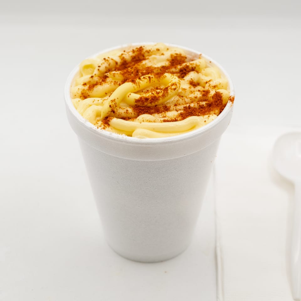 Creamy macaroni and cheese topped with reddish seasoning in a white takeout cup, with a white spoon beside it.