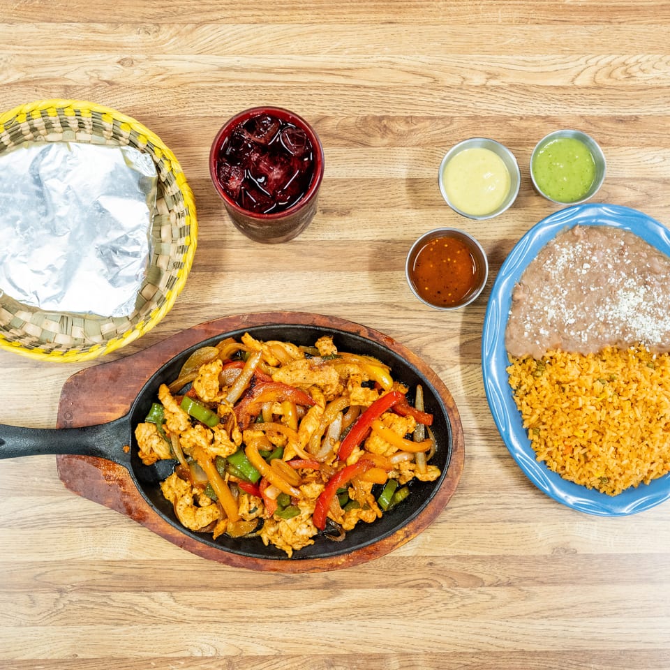 Overhead view of sizzling chicken fajitas with peppers, Mexican rice, refried beans, tortillas, sauces, and a red drink on a wooden table.